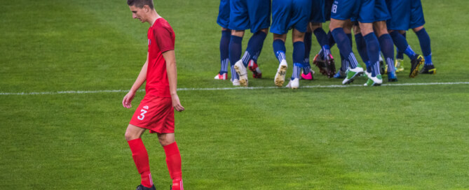 Disappointed soccer player walks across field while opposing team celebrates success in background