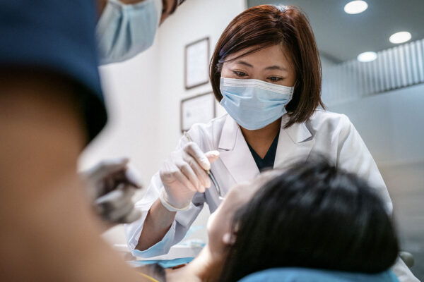 Female dentist wearing surgical mask treating woman. Male coworker is assisting mid adult healthcare worker. Medical professionals are examining patient in clinic.